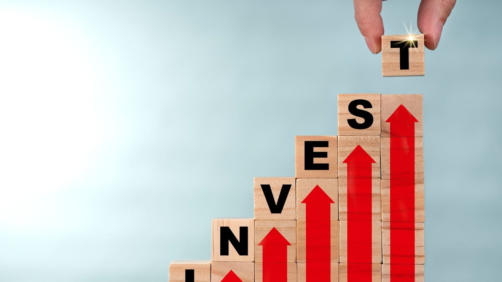 A hand placing the final wooden block on a stack with letters spelling "INVEST" and upward-trending red arrows painted on the side, symbolizing the growth and success of strategic investments — The Most Profitable Business in Canada