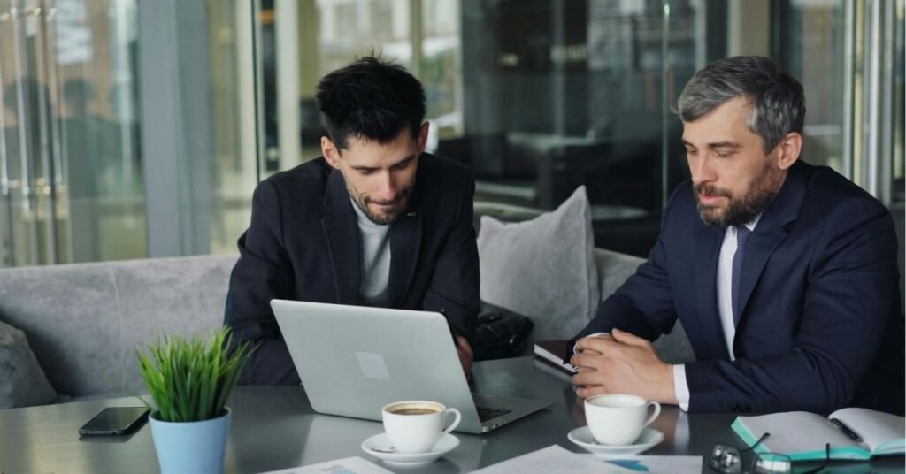 Two men in business attire sitting at a table in a modern office café, looking at a laptop together with coffee cups, documents, and a small plant on the table.