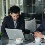 Two men in business attire sitting at a table in a modern office café, looking at a laptop together with coffee cups, documents, and a small plant on the table.