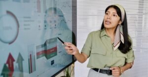A woman pointing at a large screen displaying charts and graphs during a presentation, standing in front of window blinds.