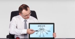 A man in a white shirt and tie sitting at a desk, holding a tablet that displays an SEO diagram labeled ‘Website’ with arrows pointing to elements like links, content, media, and rank.
