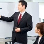 A man in a business suit giving a presentation to colleagues, pointing at charts on a whiteboard while three people seated around him listen.