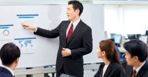 A man in a business suit giving a presentation to colleagues, pointing at charts on a whiteboard while three people seated around him listen.