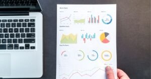 A person holding a printed sheet of various colorful charts and graphs next to an open laptop on a dark desk.