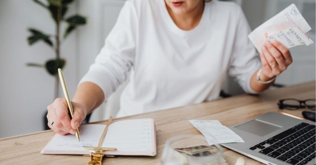Woman sitting at a desk reviewing receipts and writing in a planner, with a laptop, glasses, and other documents nearby—preparing financial records for tax season.