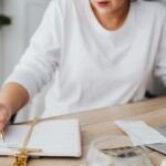 Woman sitting at a desk reviewing receipts and writing in a planner, with a laptop, glasses, and other documents nearby—preparing financial records for tax season.
