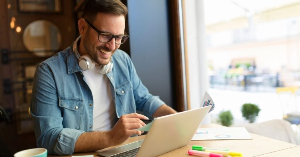 Smiling man wearing glasses and headphones around his neck working on a laptop at a bright, modern café, holding a credit card with colorful charts and highlighters on the table.