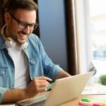 Smiling man wearing glasses and headphones around his neck working on a laptop at a bright, modern café, holding a credit card with colorful charts and highlighters on the table.