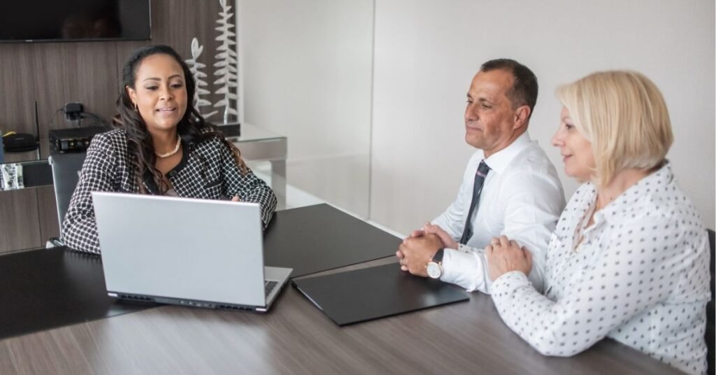 A professional woman presents information on a laptop to a mature couple seated across the table. The meeting appears formal, possibly a financial or legal consultation, with the participants attentively engaged in a modern office setting.