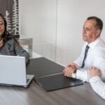 A professional woman presents information on a laptop to a mature couple seated across the table. The meeting appears formal, possibly a financial or legal consultation, with the participants attentively engaged in a modern office setting.