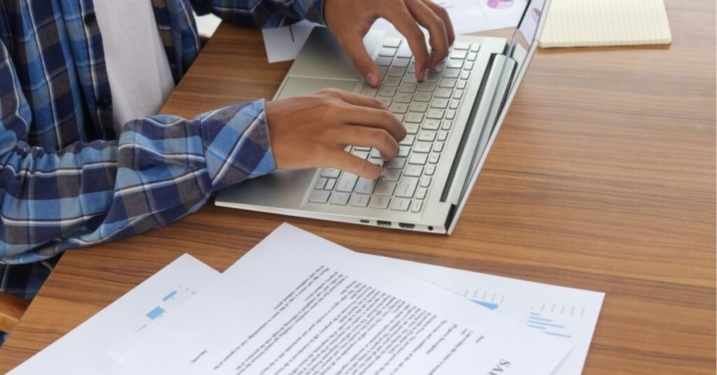 Close-up of a person typing on a silver laptop at a wooden desk, surrounded by printed documents and charts. The individual is wearing a blue plaid shirt, and the workspace appears organized with business-related papers.