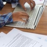 NUANS in Canada: Choose and Secure Your Business Name Close-up of a person typing on a silver laptop at a wooden desk, surrounded by printed documents and charts. The individual is wearing a blue plaid shirt, and the workspace appears organized with business-related papers.