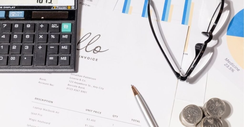 Close-up of a desk with financial documents, a calculator, a silver pen, eyeglasses, coins, and charts. An invoice addressed to Jonathan Patterson is partially visible, alongside business-related graphs and pie charts.