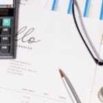 Close-up of a desk with financial documents, a calculator, a silver pen, eyeglasses, coins, and charts. An invoice addressed to Jonathan Patterson is partially visible, alongside business-related graphs and pie charts.