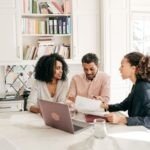 A financial advisor meets with a couple at their kitchen table, discussing documents. The advisor, dressed in business attire, gestures toward a paper while the couple listens attentively. A laptop, notebooks, and water glasses are on the table, with a bright kitchen and bookshelf in the background.